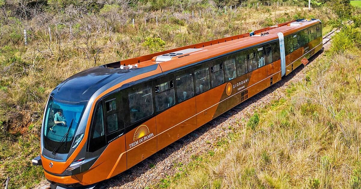 Imagem de um trem moderno conhecido como Trem do Pampa, percorrendo as planícies verdes do Rio Grande do Sul, com vista panorâmica e céu azul ao fundo.