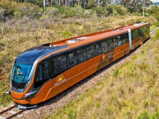 Imagem de um trem moderno conhecido como Trem do Pampa, percorrendo as planícies verdes do Rio Grande do Sul, com vista panorâmica e céu azul ao fundo.