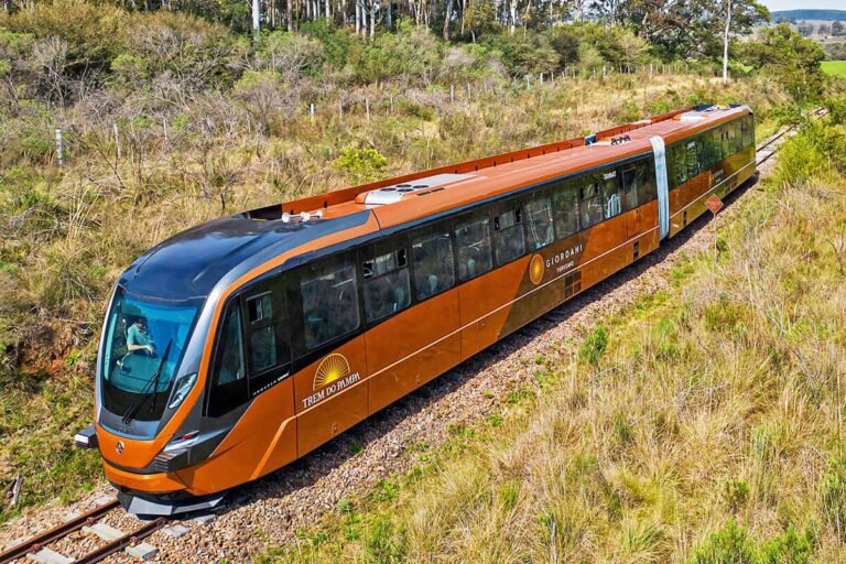 Imagem de um trem moderno conhecido como Trem do Pampa, percorrendo as planícies verdes do Rio Grande do Sul, com vista panorâmica e céu azul ao fundo.